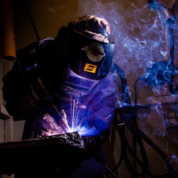 Students leans over their welding. They're wearing full safety equipment, and their work is producing purple sparks and smoke in the air. It looks very dramatic yet atmospheric. 