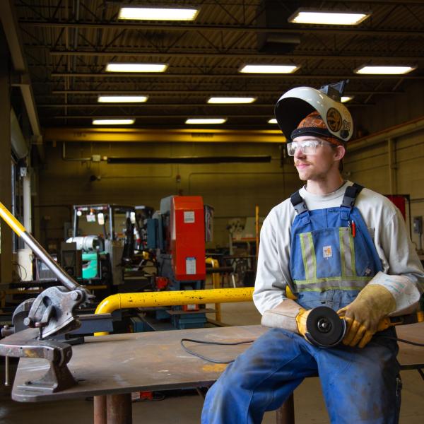 A welding student in full protective gear sits on a work bench and looks into the distance. 