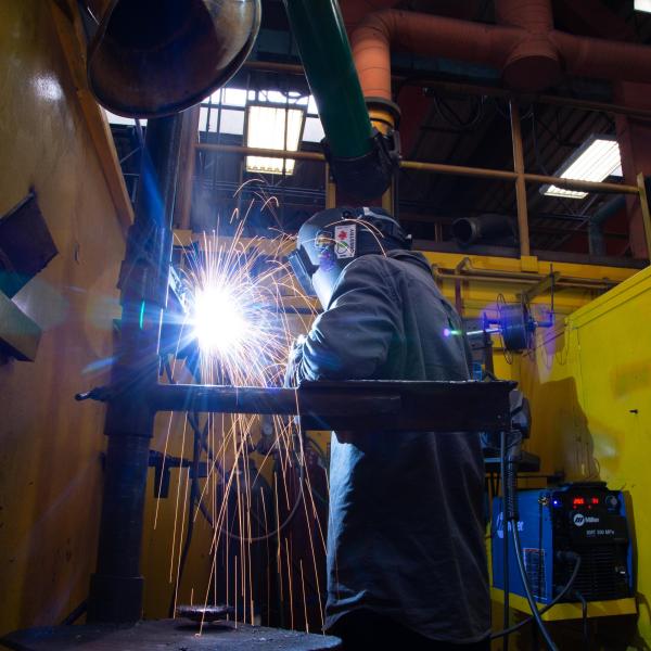 A VIU welding student is practicing their skills in a welding booth. The booth walls are yellow, the student's wearing blue, and the sparks from the welding are bright orange. 