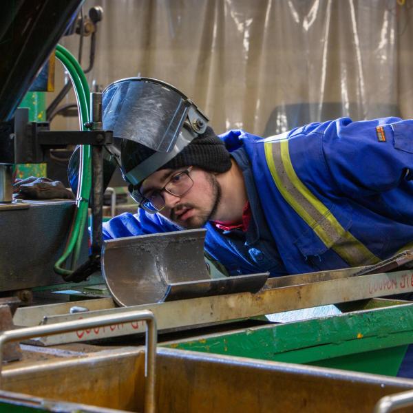 A view welding student has pushed up his protective head gear to look closely at their work. 