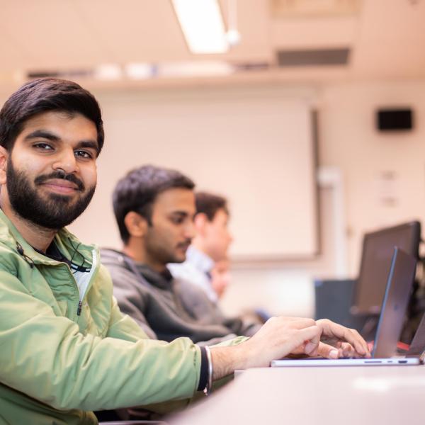 We are looking down a row of VIU ITAS students who are working on their laptops. The student in the foregound is smiling and looking directly at the camera with his hands on his keyboard. 