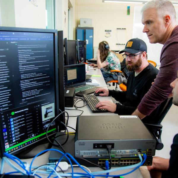 A row of VIU ITAS students are sat in front vertical computer monitors. An instructor stands amongst them and is guiding their work. 