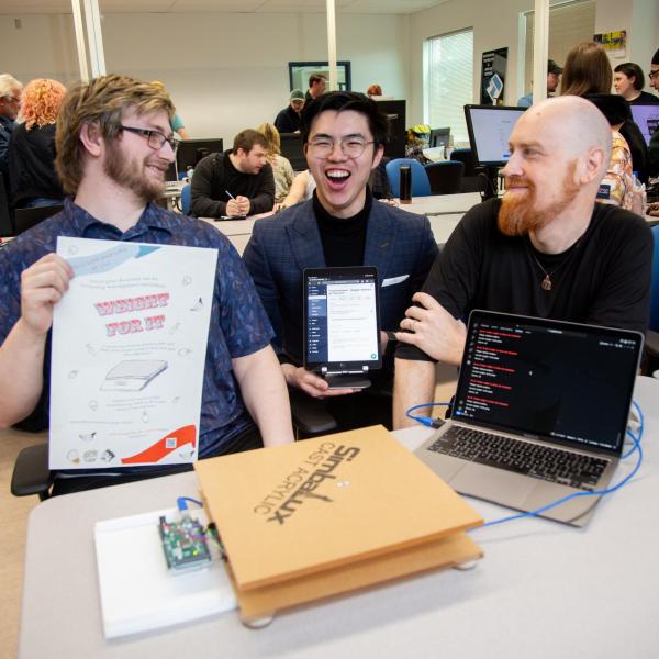 A group of three VIU ITAS students sit side by side. They are holding up parts of their final project. The person in the middle is laughing and the other two are looking at him smiling. 