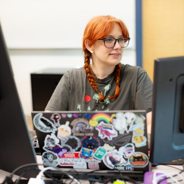 A female VIU ITAS student is sitting in front of her laptop which is flanked on each side with a large monitor. Her laptop is covered in stickers and full of personality. 