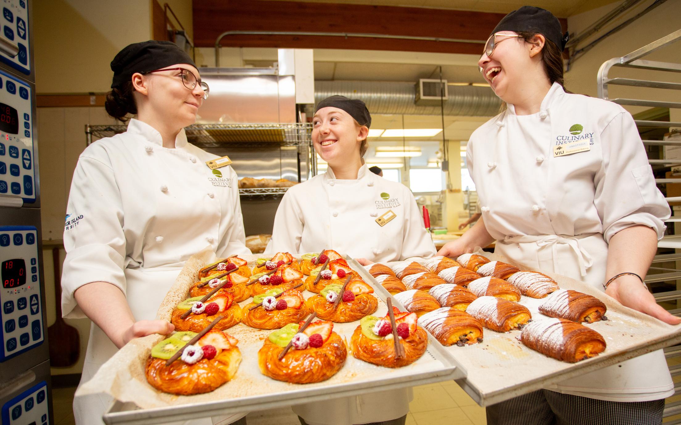 Three women in chef's whites with trays of baked goods. 