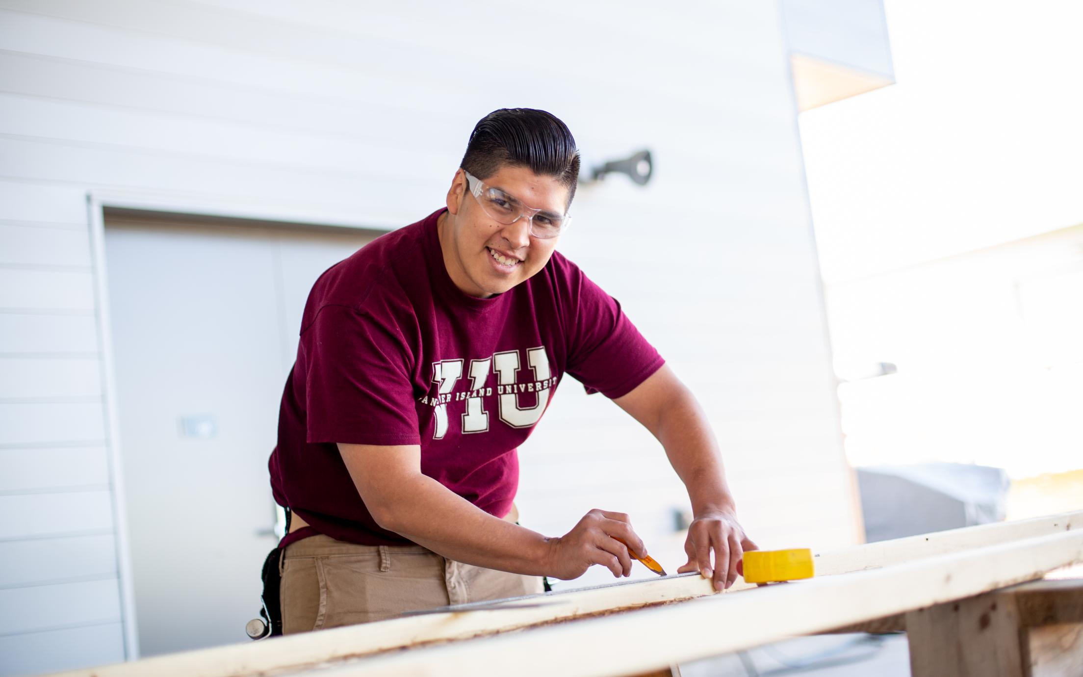 Indigenous man in red VIU shirt measuring a piece of wood in carpentry class, smiling