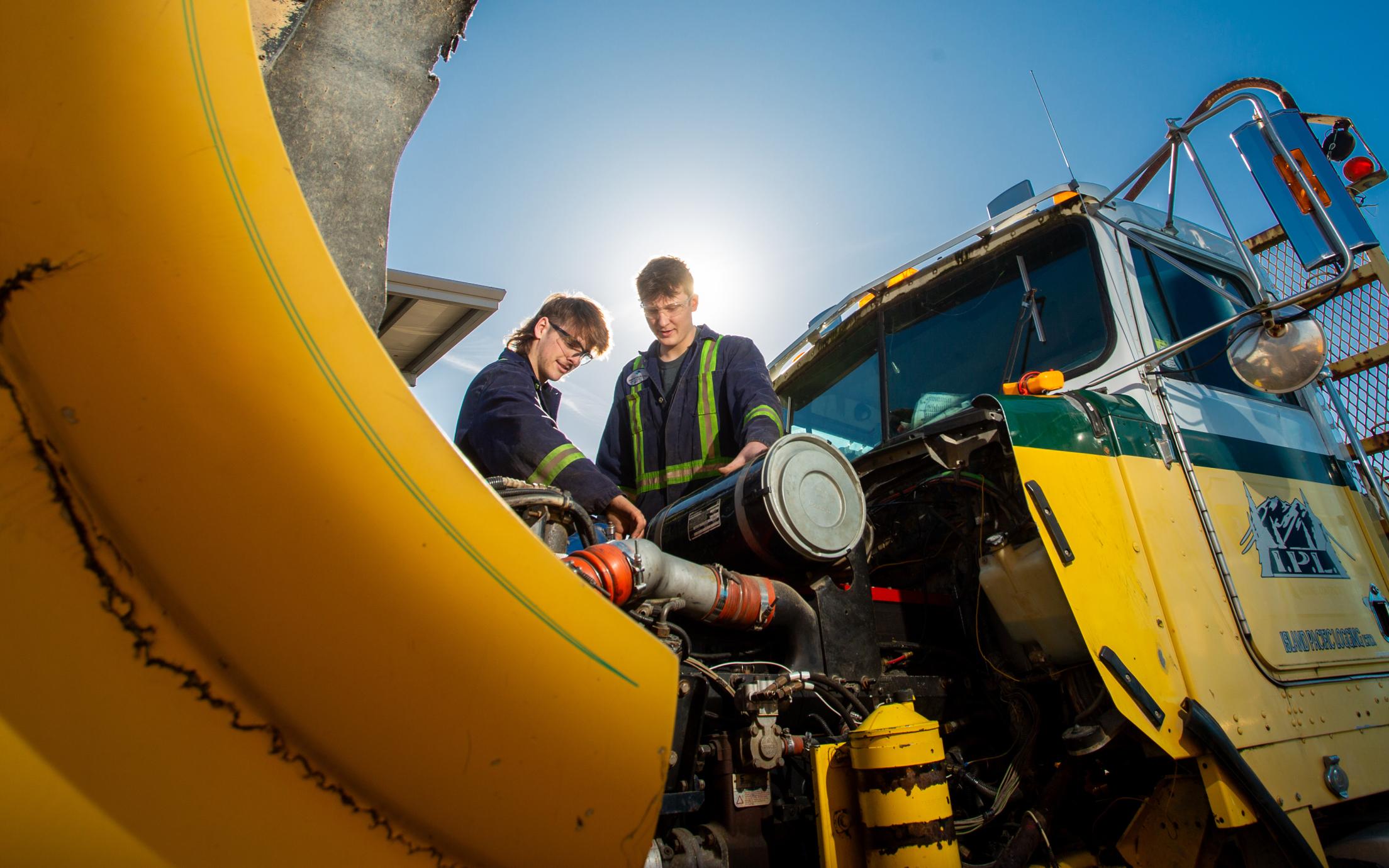 Two men working on the engine of a big yellow truck under a sunny blue sky. 